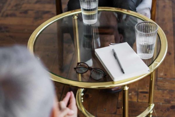 Small glass table with notepad and pen on it and two glasses of water and two people either side in the background
